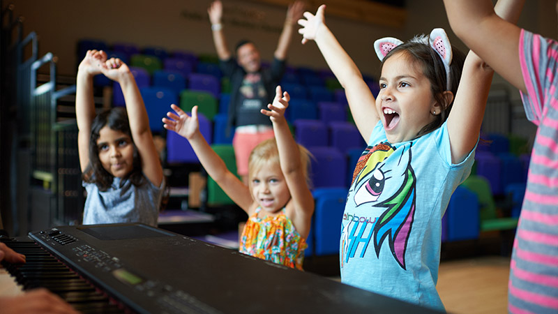 Children playing piano in an auditorium: A group of kids joyfully playing the piano together in a large auditorium.